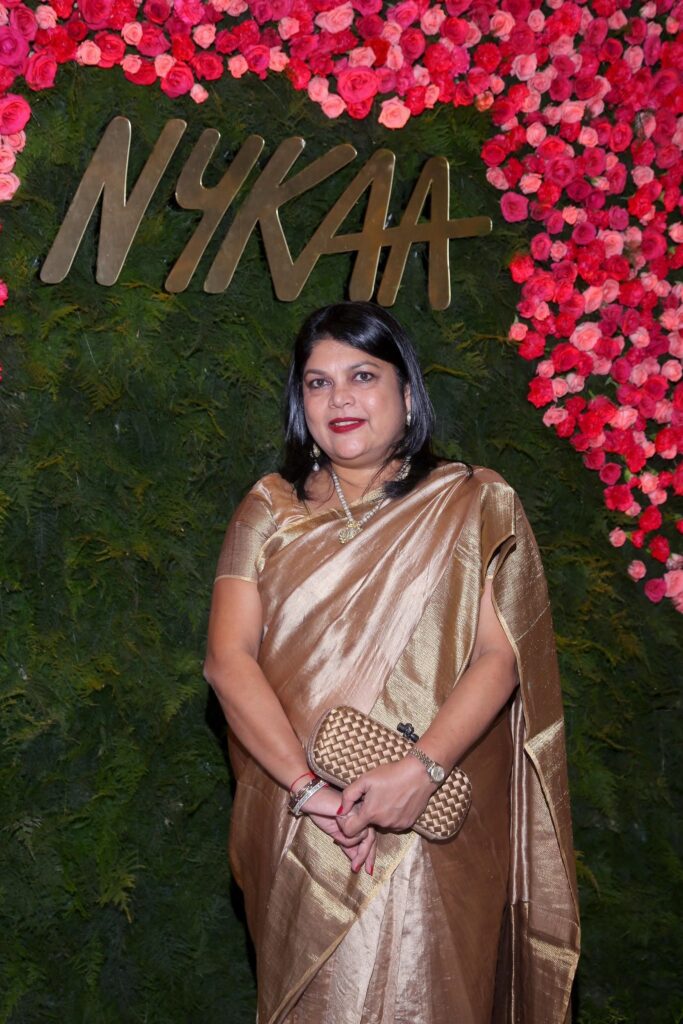 A woman in a golden saree posing at a Nykaa event, standing against a floral and greenery backdrop with the Nykaa logo.