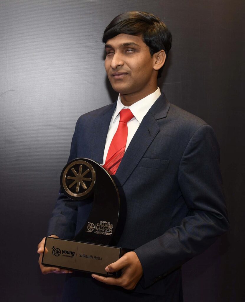 Srikanth Bolla in a suit holding an award trophy, standing against a black background.