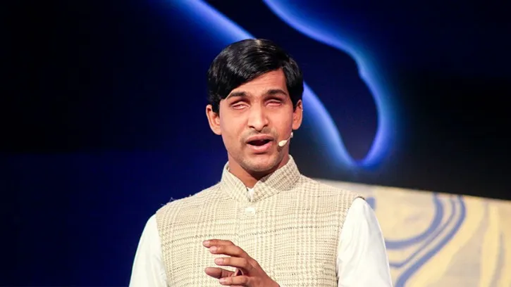 Srikanth Bolla speaking on stage, dressed in a white kurta with a microphone headset.