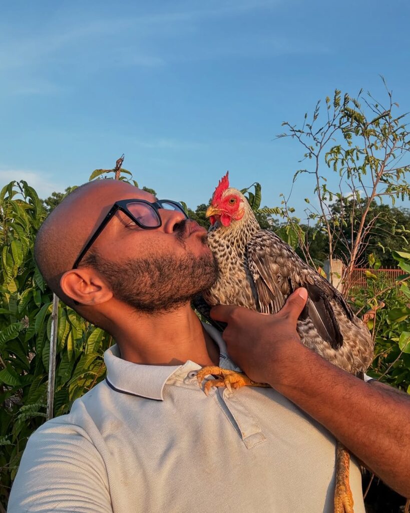 A man wearing glasses holds a chicken close to his face while standing outdoors under clear blue skies.