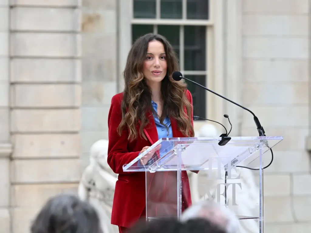 Woman in a red blazer speaking at a podium with microphones.