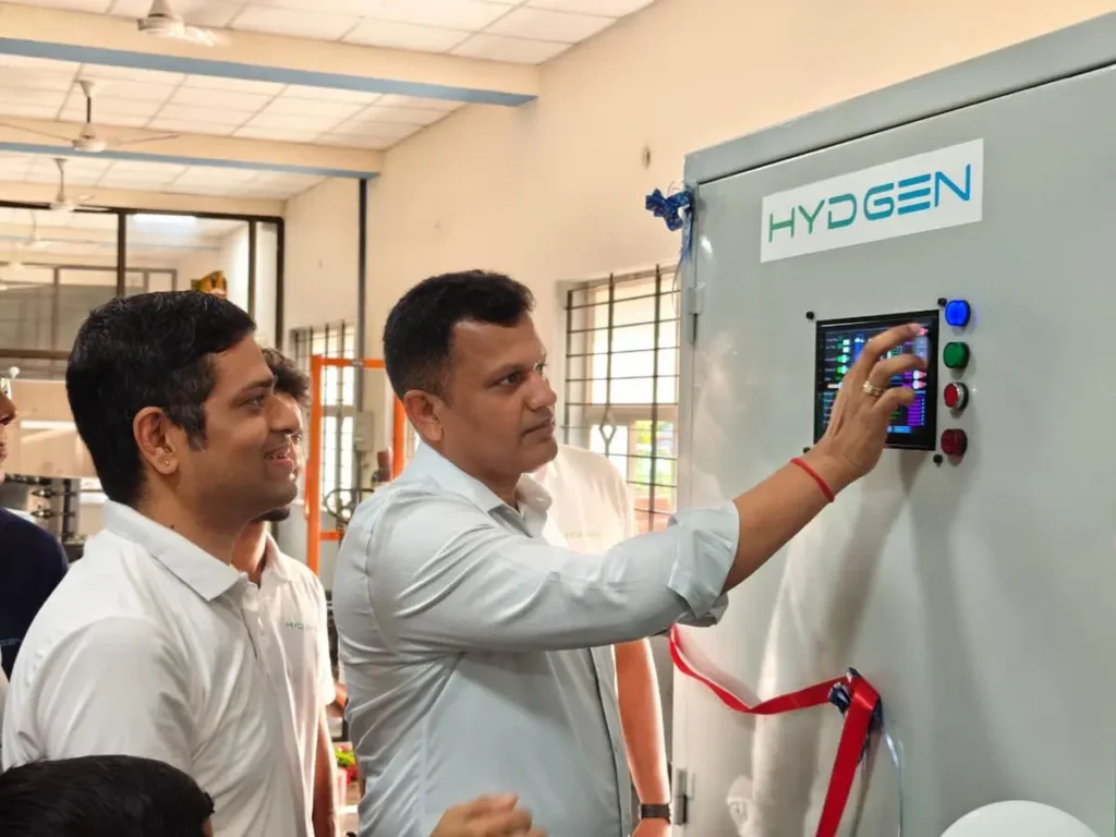 Two HYDGEN engineers press controls on a HYDGEN-branded cabinet containing a hydrogen electrolyzer, with a red ribbon attached at its base indicating an inauguration event.