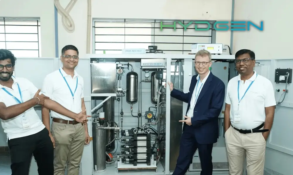 Four HYDGEN team members stand beside an open hydrogen electrolyzer cabinet inside a laboratory, pointing to its internal components, with the HYDGEN logo visible on the wall.