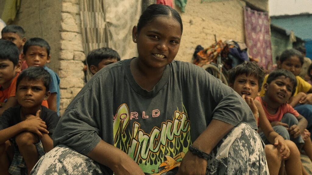 Paramjeet Kaur, the 19-year-old Punjabi singer of That Girl, sitting outdoors in her village surrounded by local children, wearing a casual oversized T-shirt and smiling confidently.